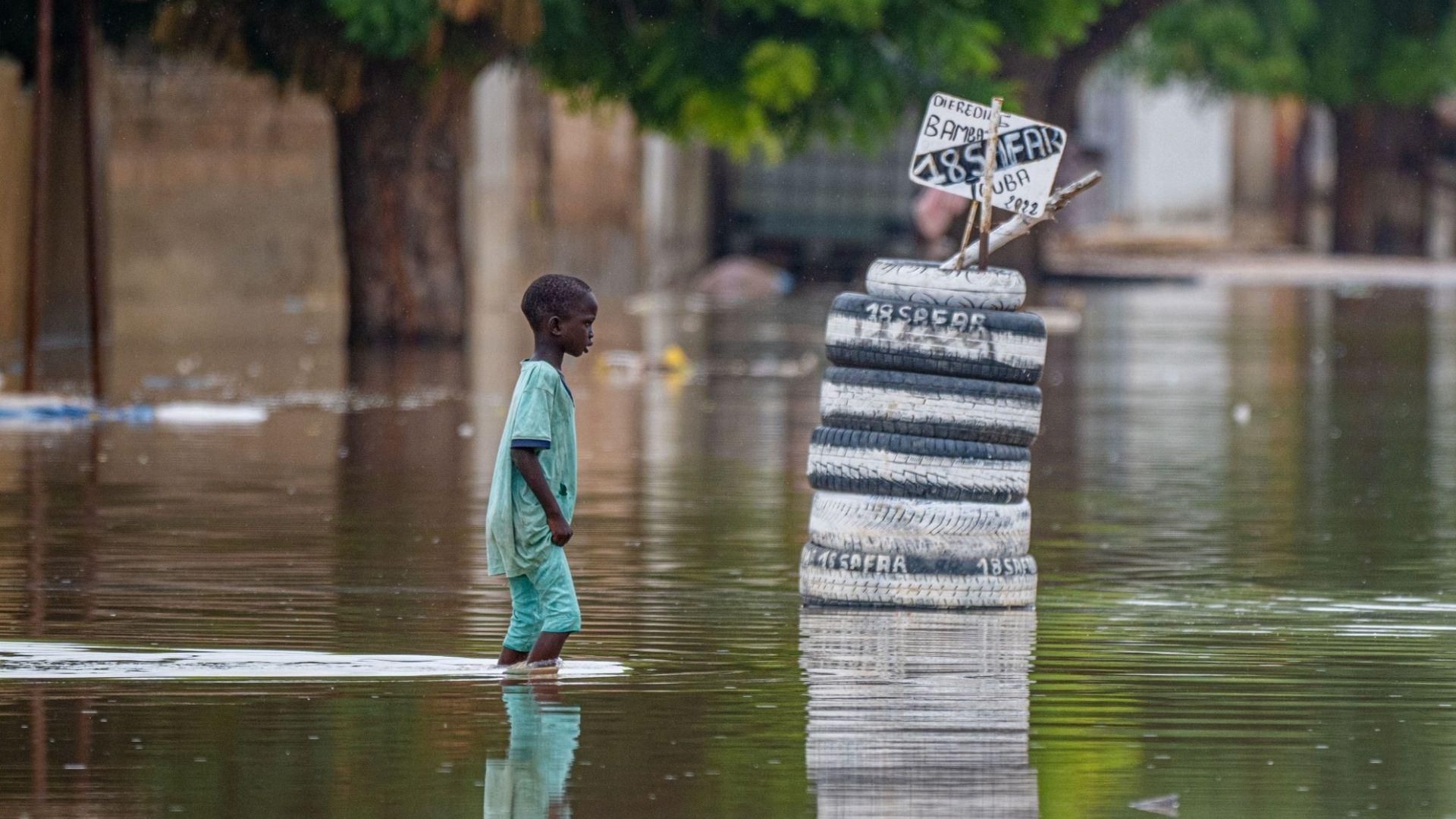 A young boy in a blue shirt standing in a flooded urban area with waist-deep water, near a stack of tires with automotive service signs