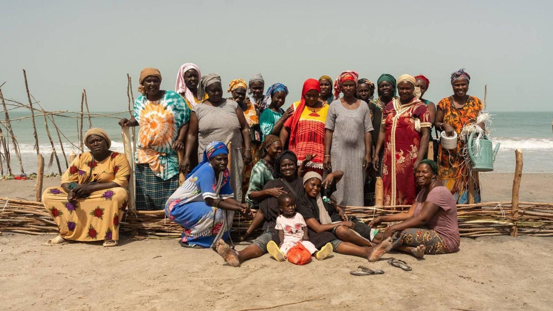 Vingt femmes et un petit enfant sourient à la caméra tout en se tenant derrière une barrière basse en palmes tressées renforcée avec des piquets en bois et de la ficelle. La barrière fait environ 50 cm de hauteur et s'étend à travers le sable avec la mer en arrière-plan.